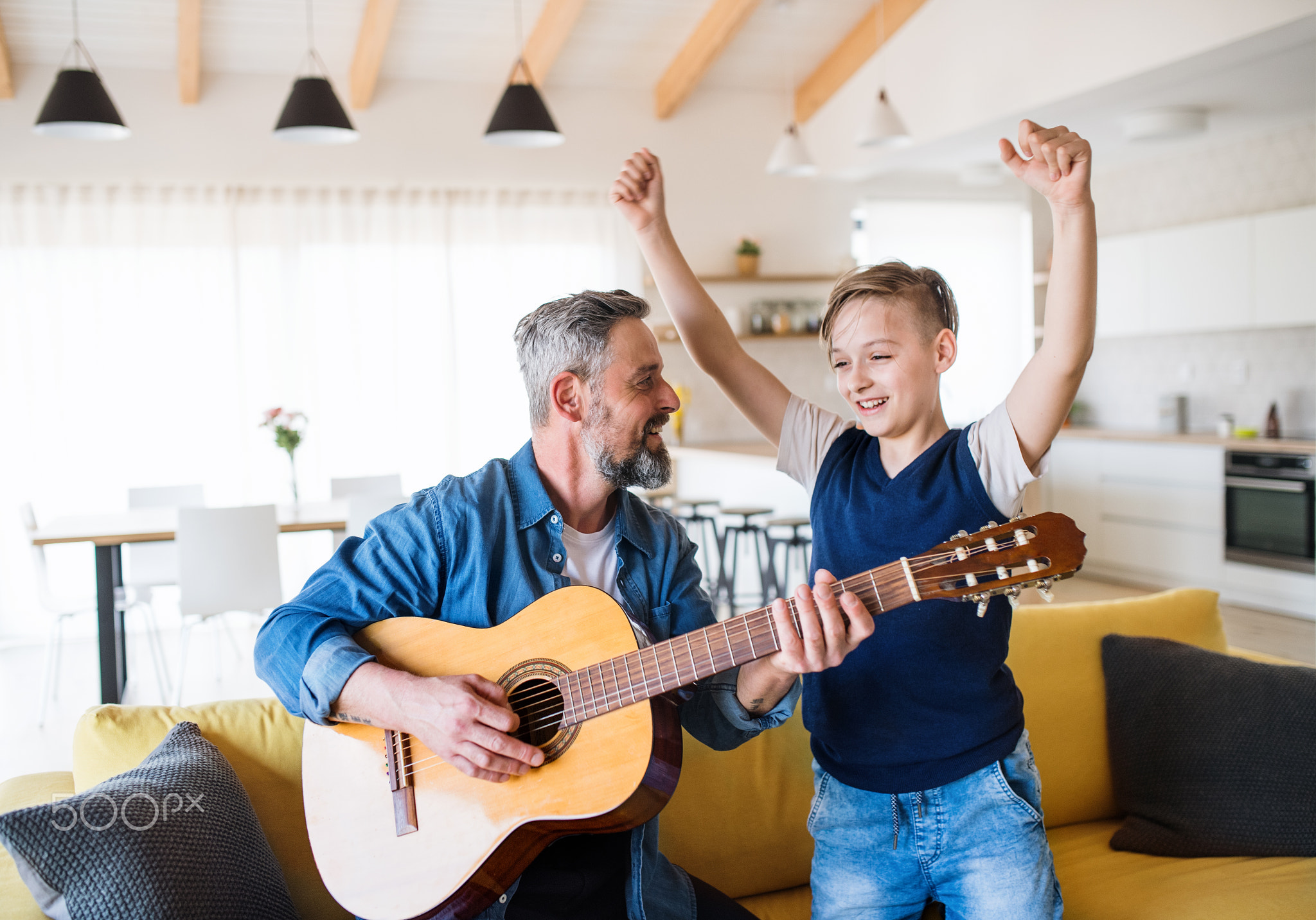Mature father with small son sitting on sofa indoors, playing guitar.