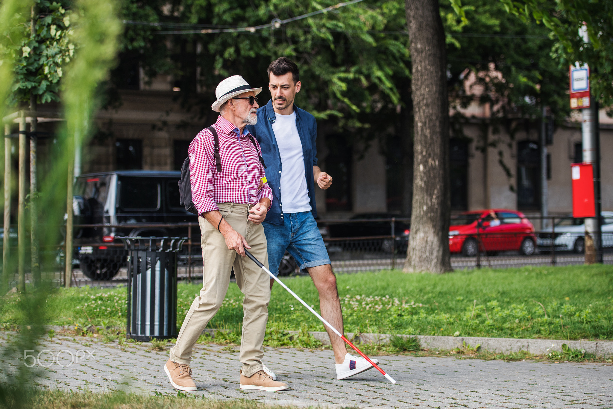 Young man and blind senior with white cane walking on pavement in city.