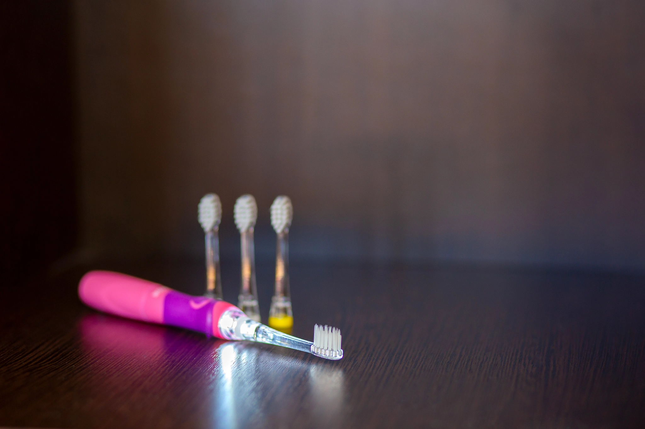 A backlit pink electric battery-powered toothbrush with three interchangeable nozzles lies on a shel