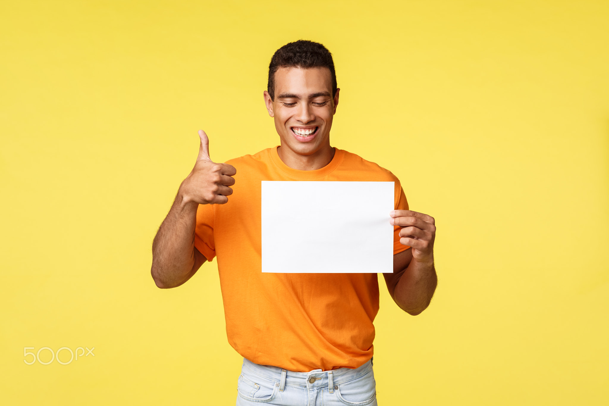 Happy handsome hispanic guy in orange t-shirt, hold blank paper, look at piece and smiling in approv