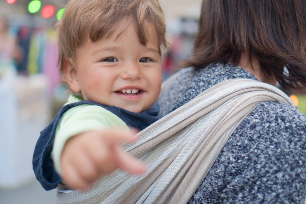 Child smiling pointing at camera carried by mom on back in baby wrap by Tamara Savkovic on 500px.com