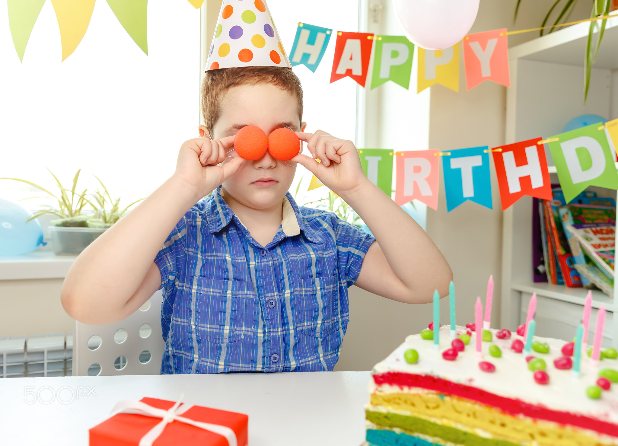 The boy put the balloons to his eyes. Birthday party and cake at home