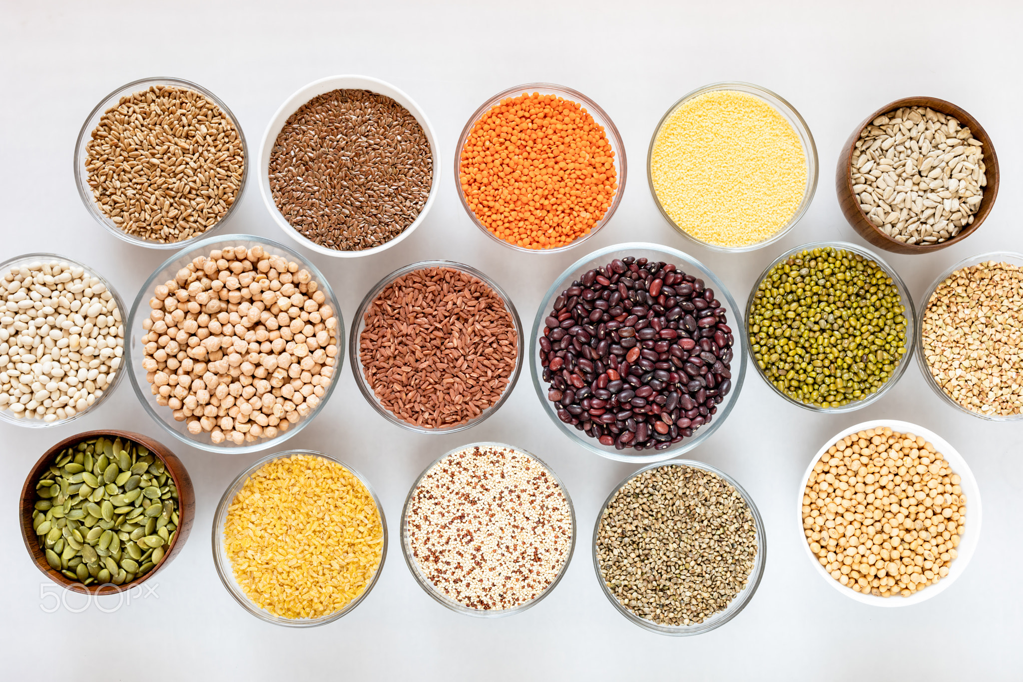 Top view to set of glass bowls with cereals, beans and seeds on white.