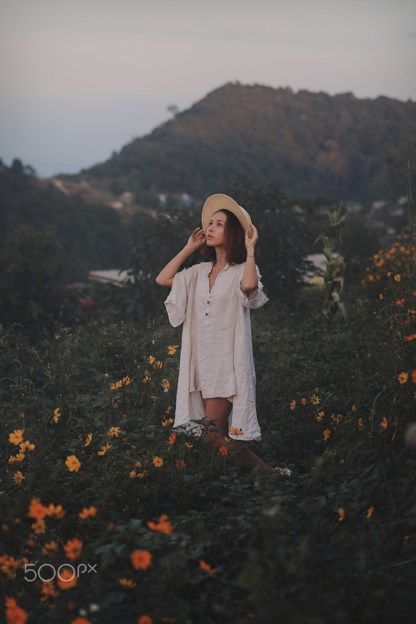 young woman in a linen dress and straw hat sits on a viewpoint in the