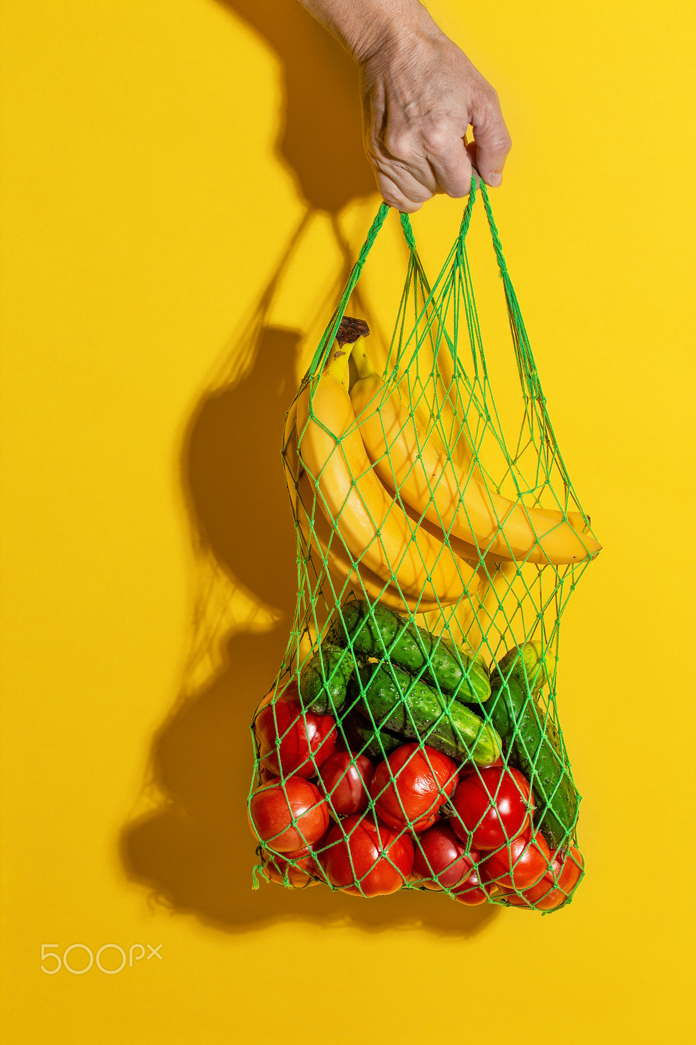 Male hand holding a white mesh bag with vegetables on yellow