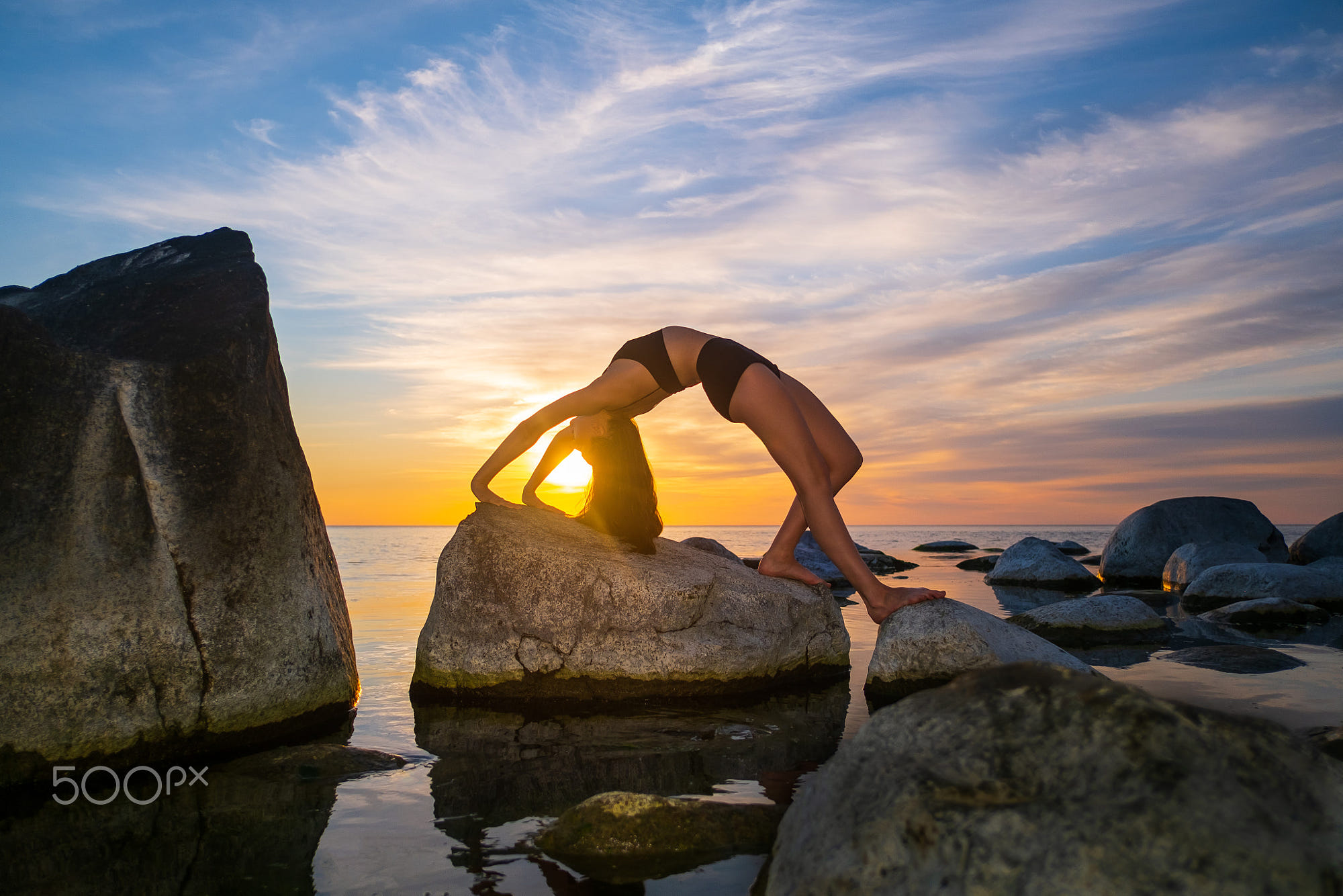 Young woman doing Wheel pose near sea: Side view of flexible female in underwear doing Urdhva Dhanurasana pose on boulders against sea and sundown sky
