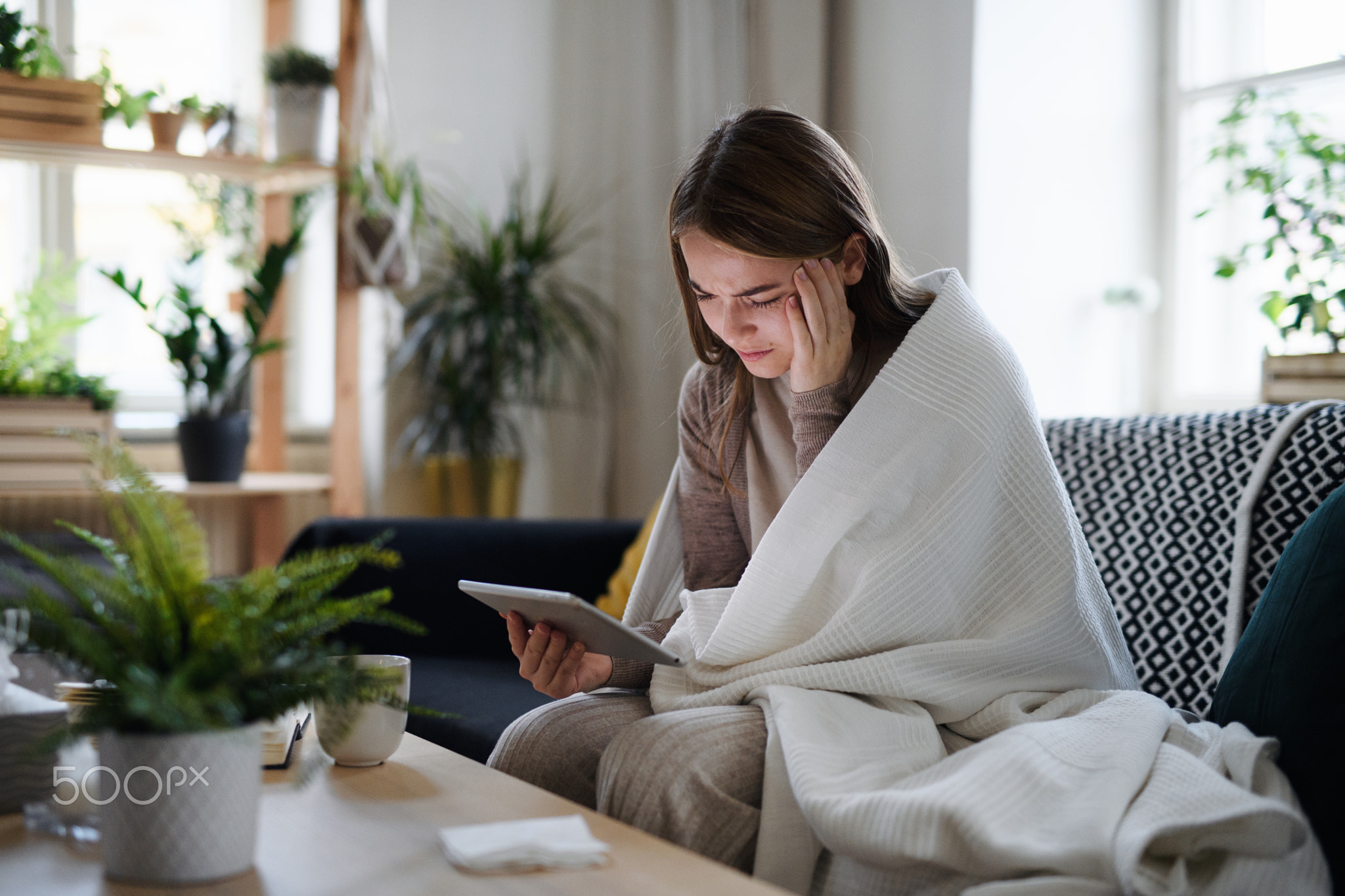 Upset young woman wrapped in blanket on sofa at home, using tablet.