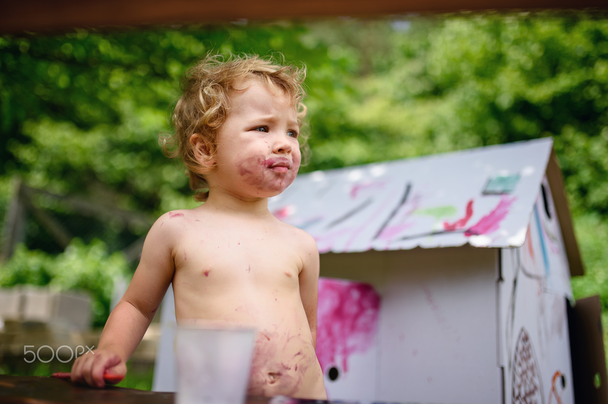 Topless small blond girl painting paper house outdoors in summer.