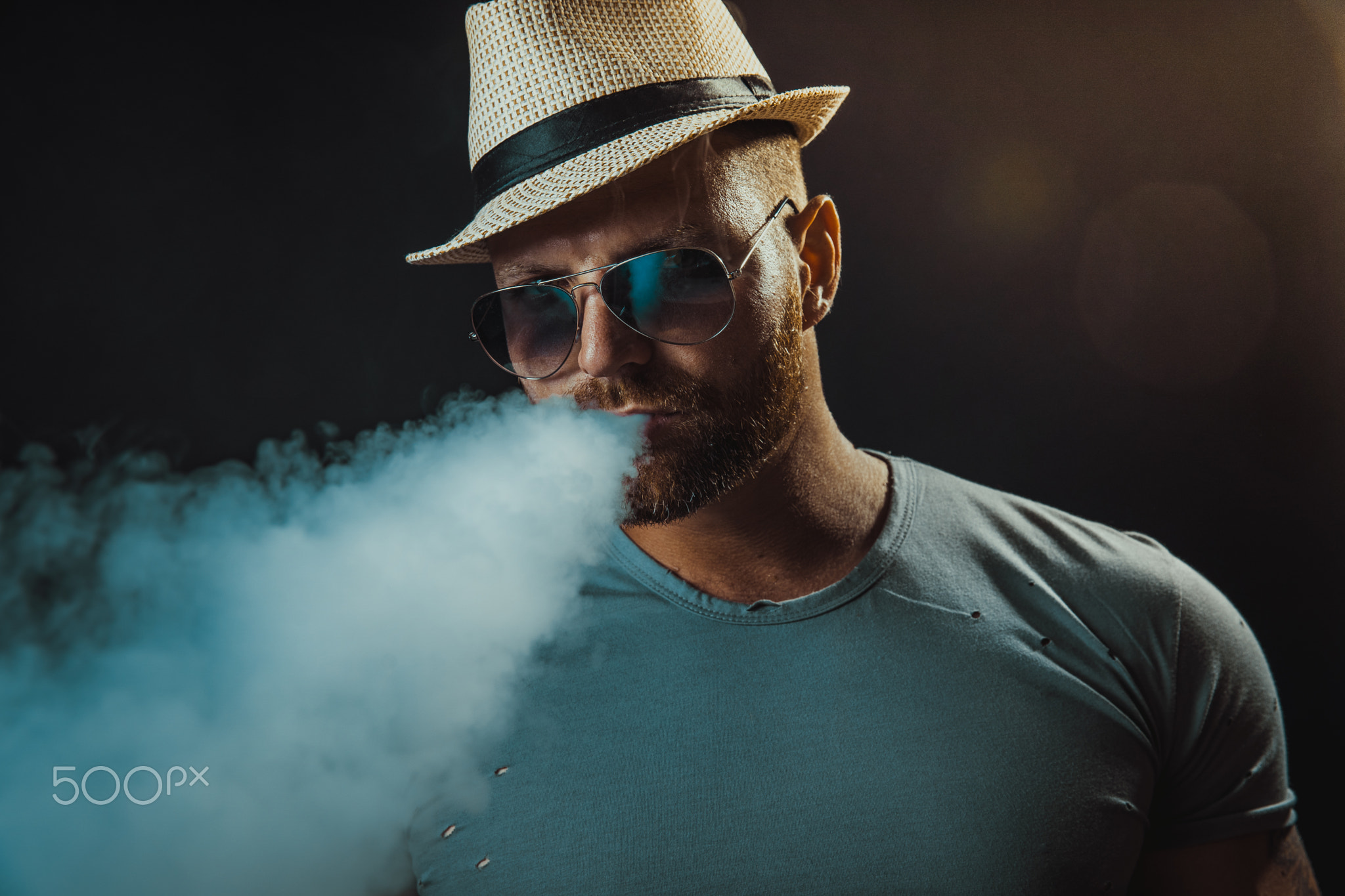 Bearded brutal male in hat and sunglasses smoking a vapor cigarette as