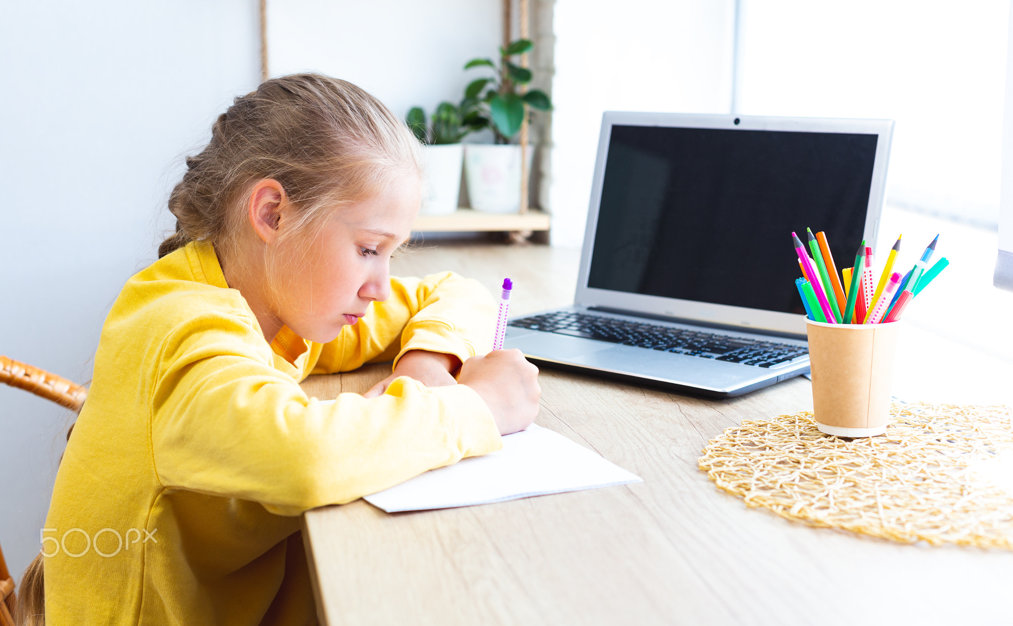 Schoolgirl in a yellow sweatshirt is doing homework at home by the