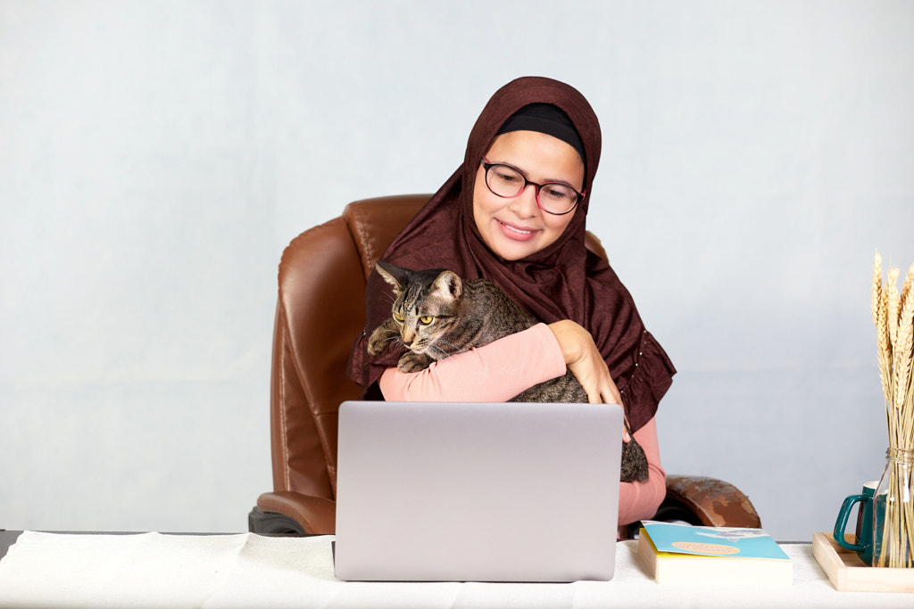 Asyalı Müslüman kadın dizüstü bilgisayarda çalışıyor ve 500px.com'da Anucha Muphasa tarafından kedisine sarılıyor.