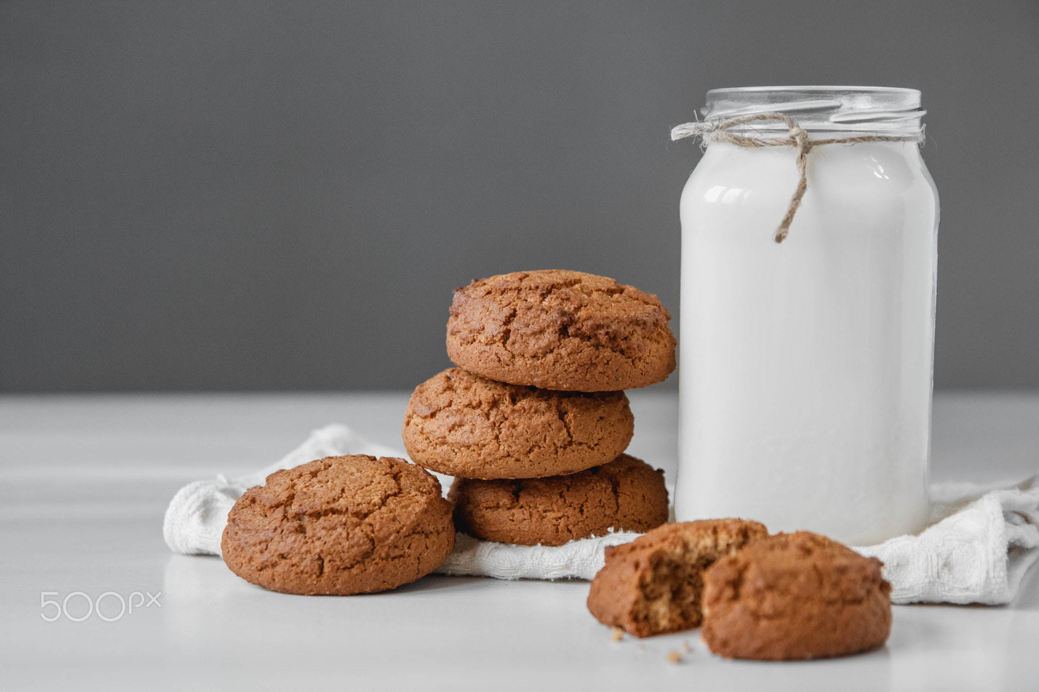 Milk in glass jar and oatmeal cookies near napkin on white table