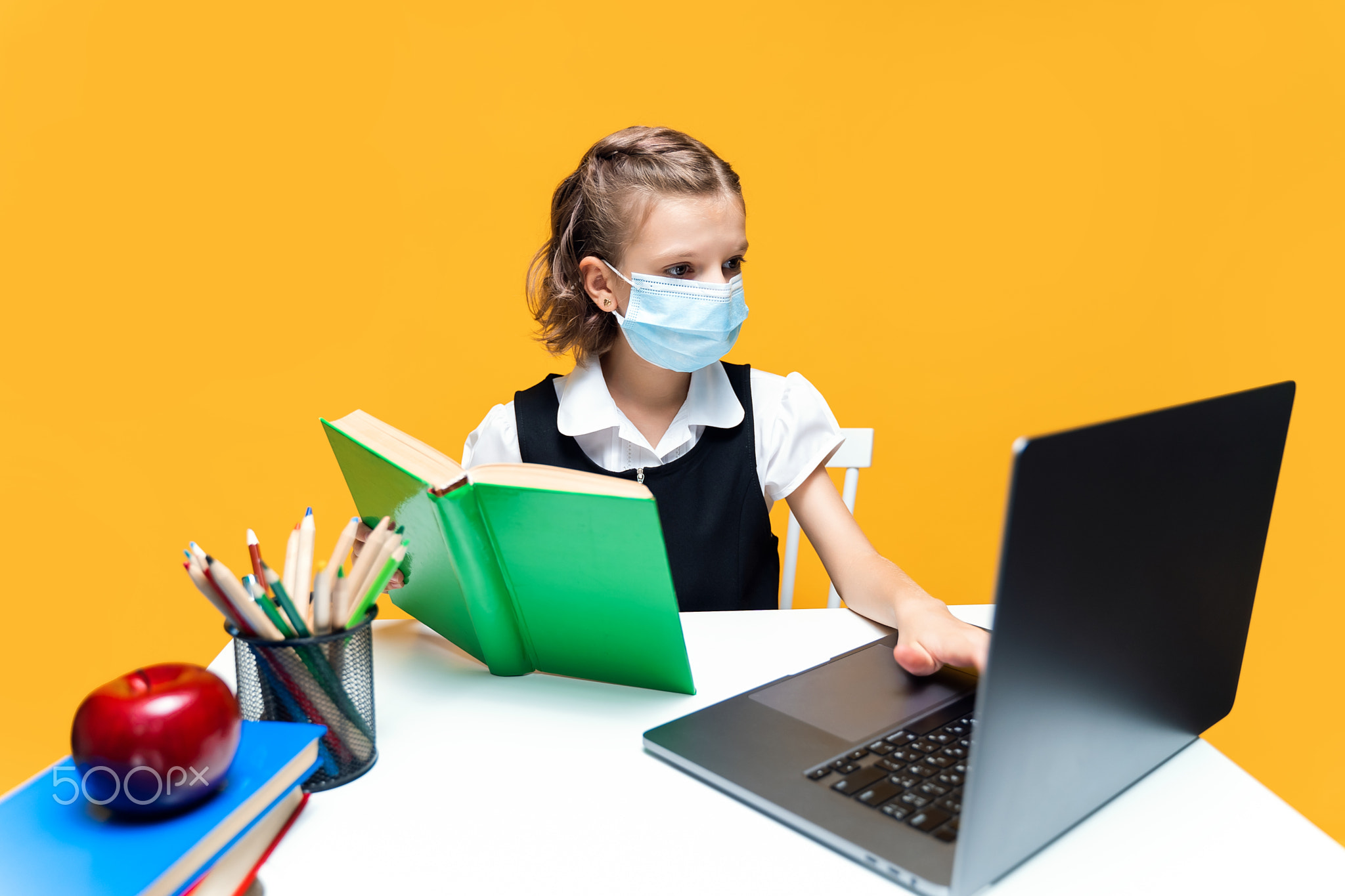 Serious schoolgirl in mask doing her homework with book and laptop