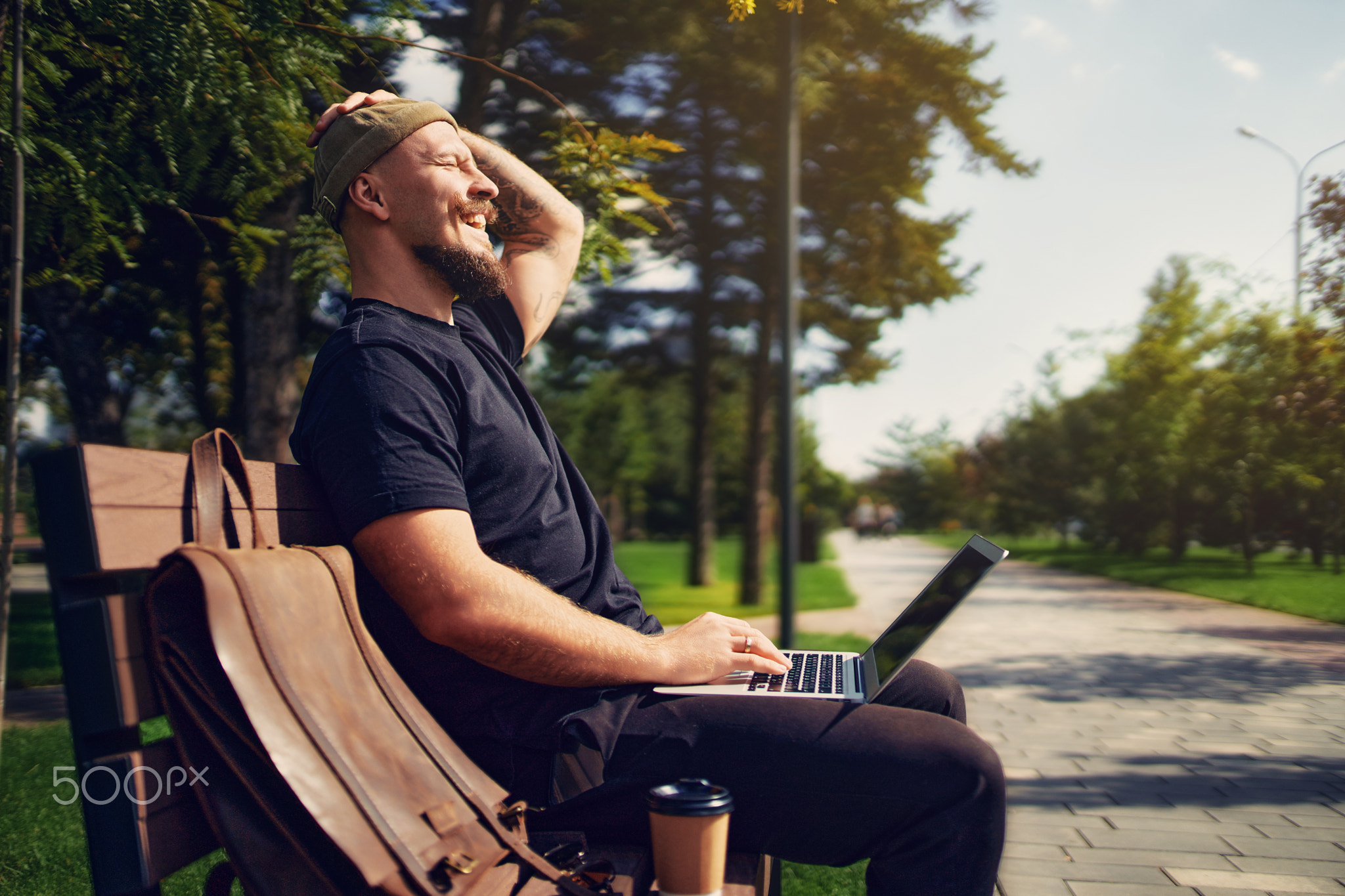 Positive guy with laptop doing remote work outside while sits on bench