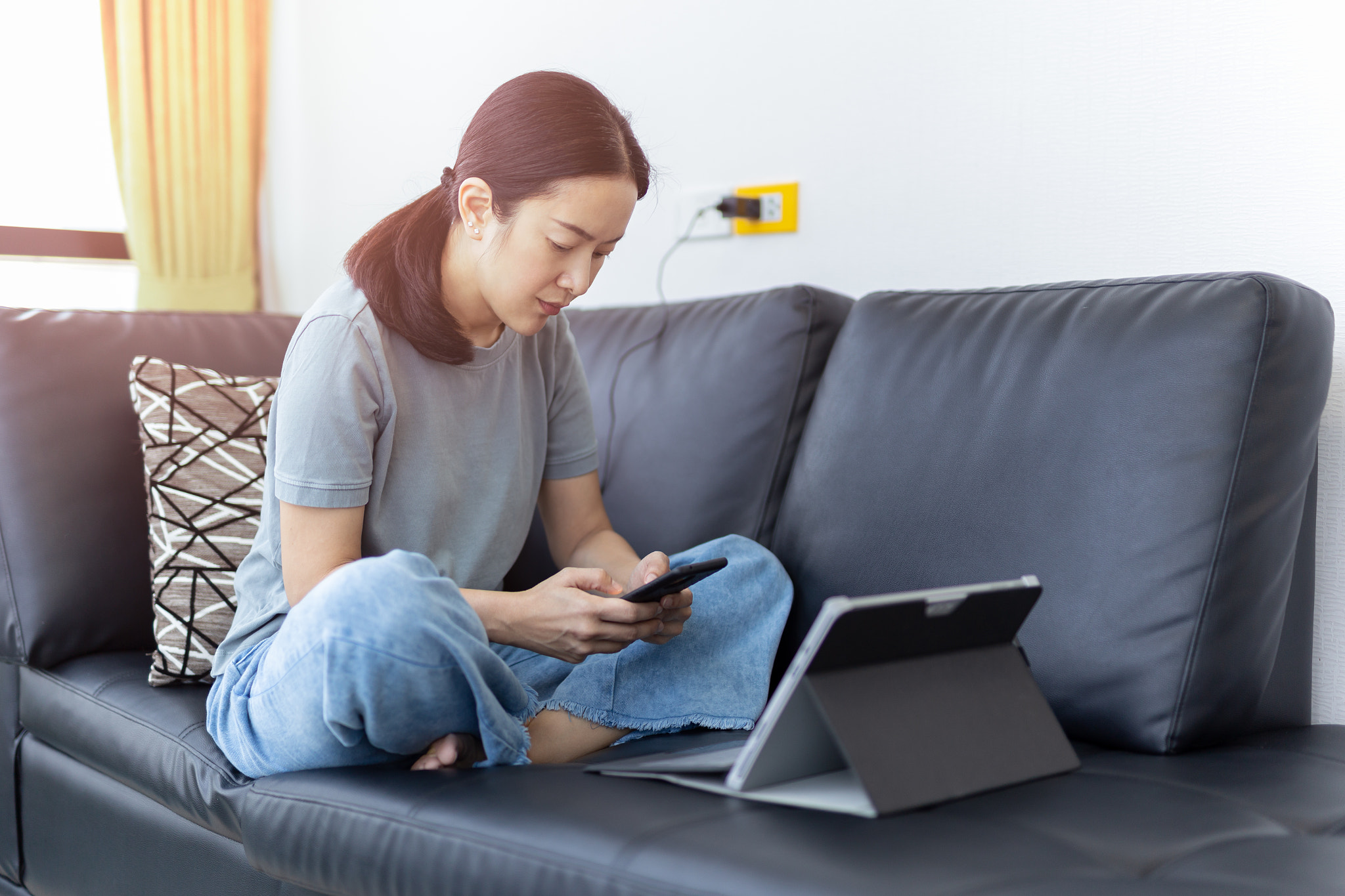 Woman using mobile phone and laptop while sitting on couch at home.