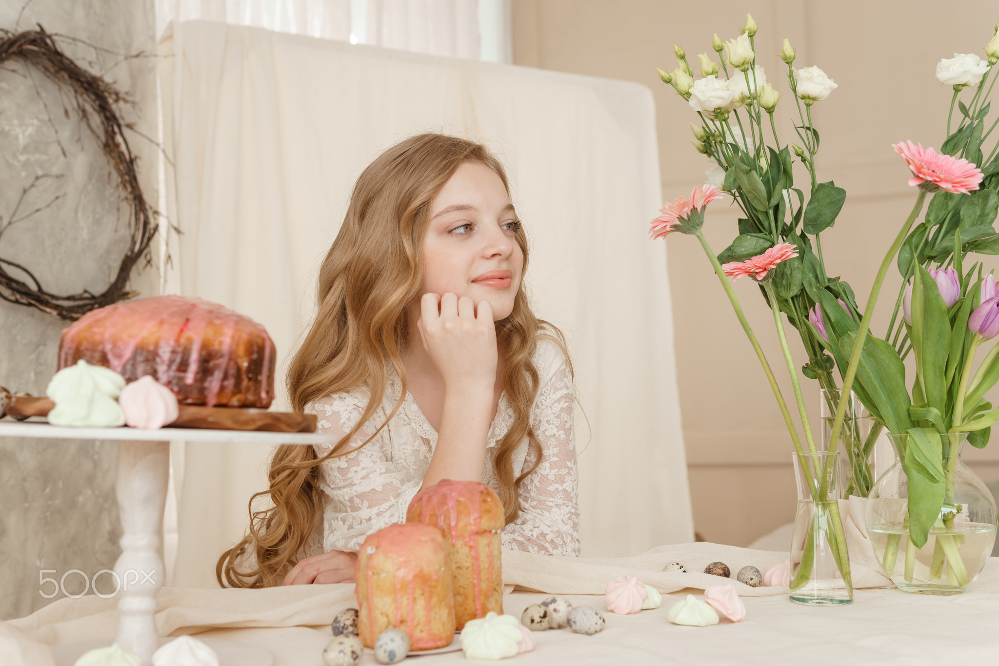 A girl with long hair in a light dress is sitting at the Easter table