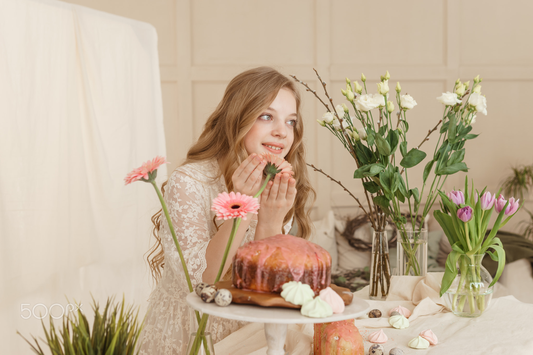 A girl with long hair in a light dress is sitting at the Easter table