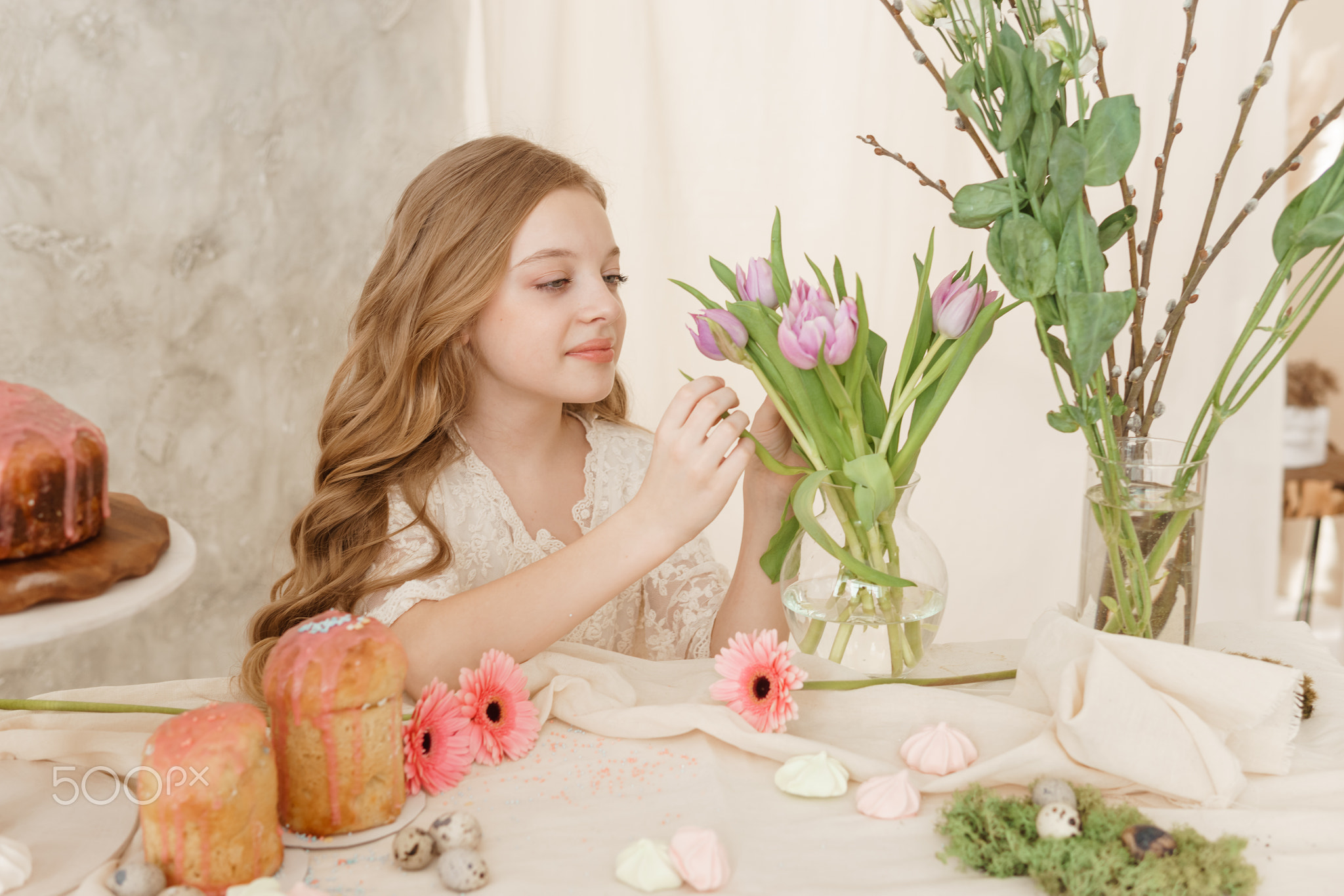 A girl with long hair in a light dress is sitting at the Easter table