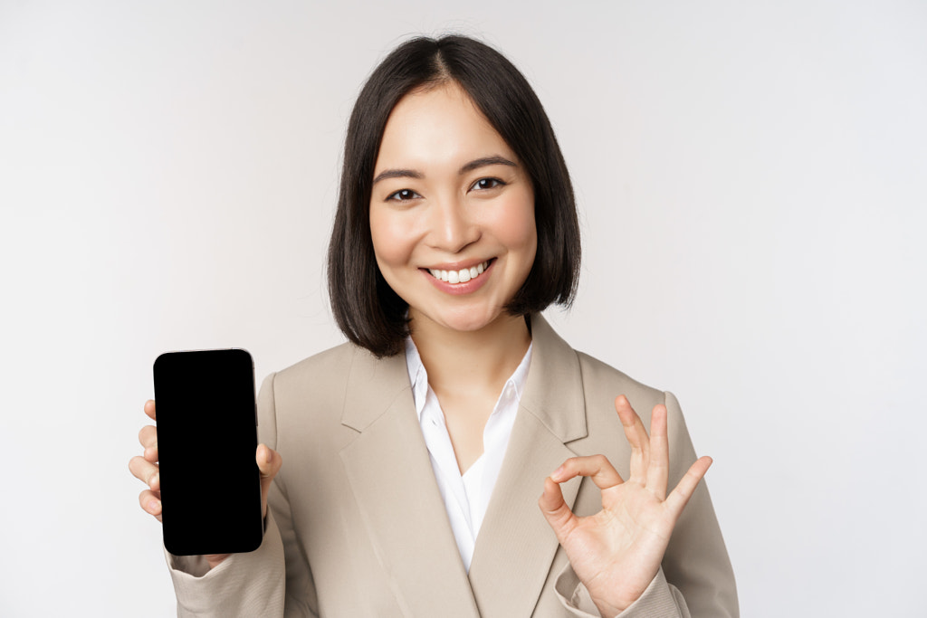 Femme asiatique souriante montrant l'écran du smartphone et le signe d'accord.  Entreprise par Mix and Match Studio sur 500px.com
