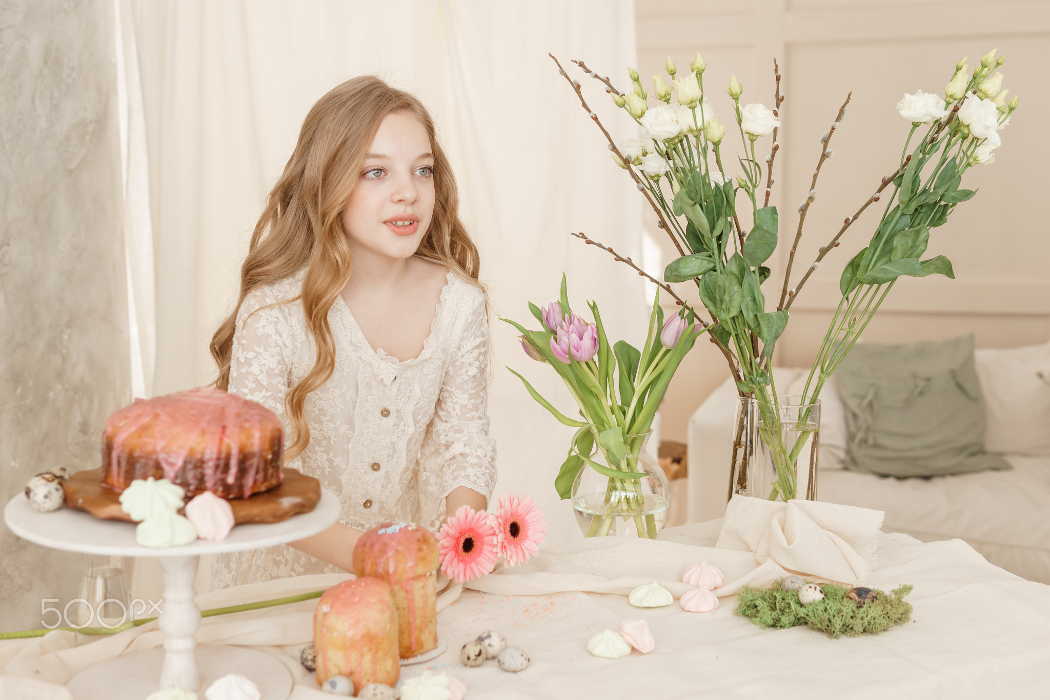 A girl with long hair in a light dress is sitting at the Easter table
