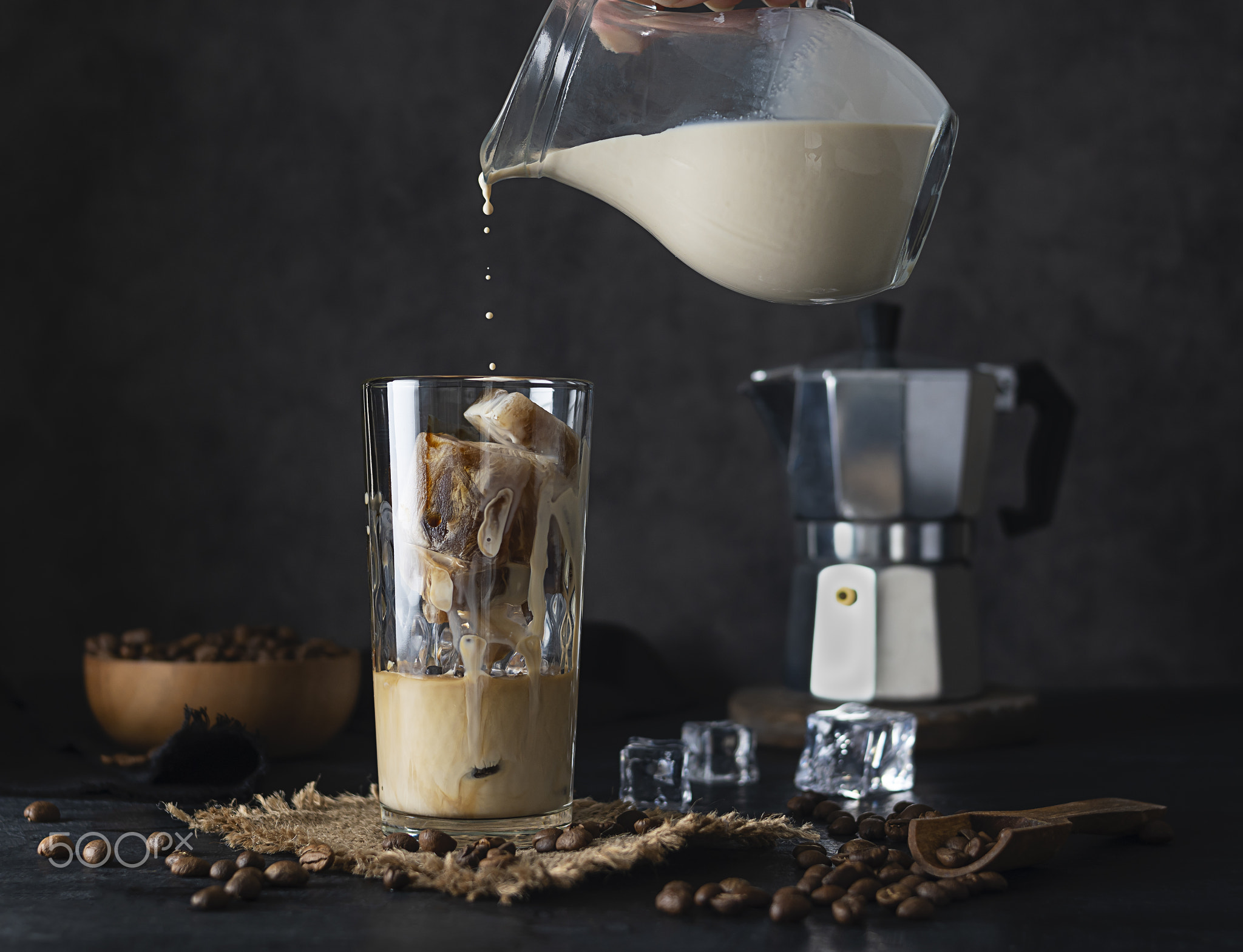 Milk from a jug pours into a transparent tall glass with ice.