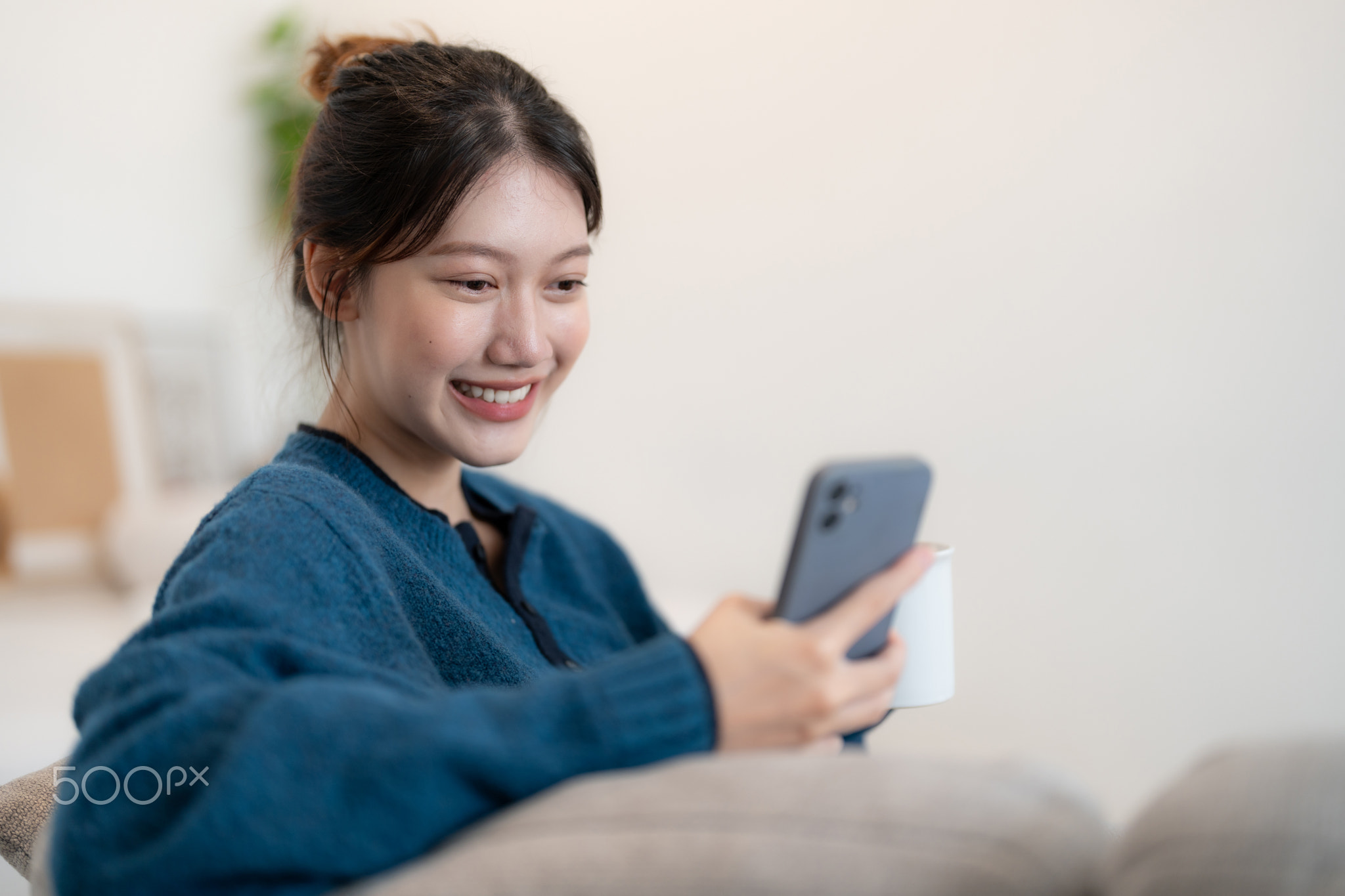 Happy young asian woman using mobile phone while sitting a couch at