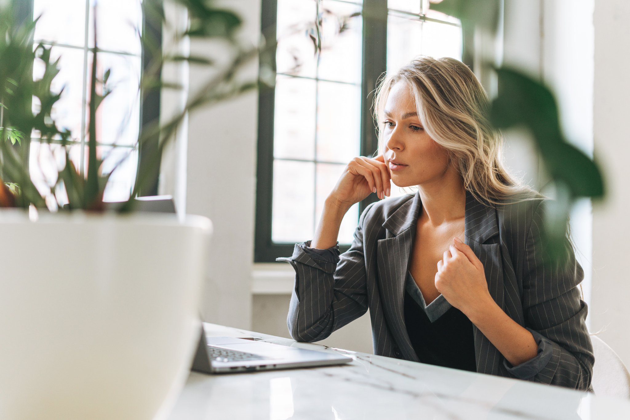 Young smiling blonde woman with long hair in stylish suit working at laptop in bright modern office