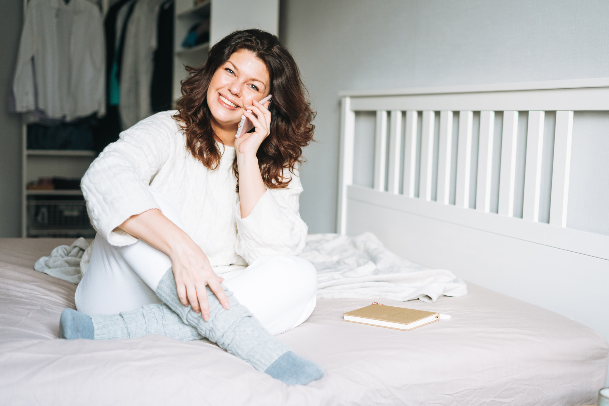 Young adult woman plus size body positive in white knitted sweater using mobile phone on bed at home