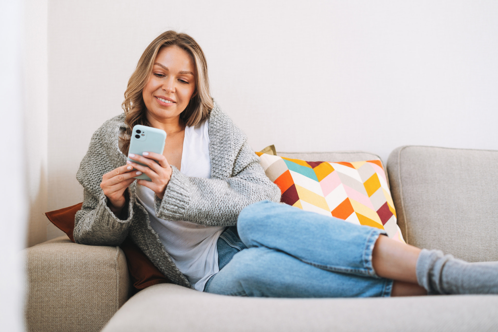 Young woman in cozy knitted grey sweater with mobile phone in hands sitting on couch at home