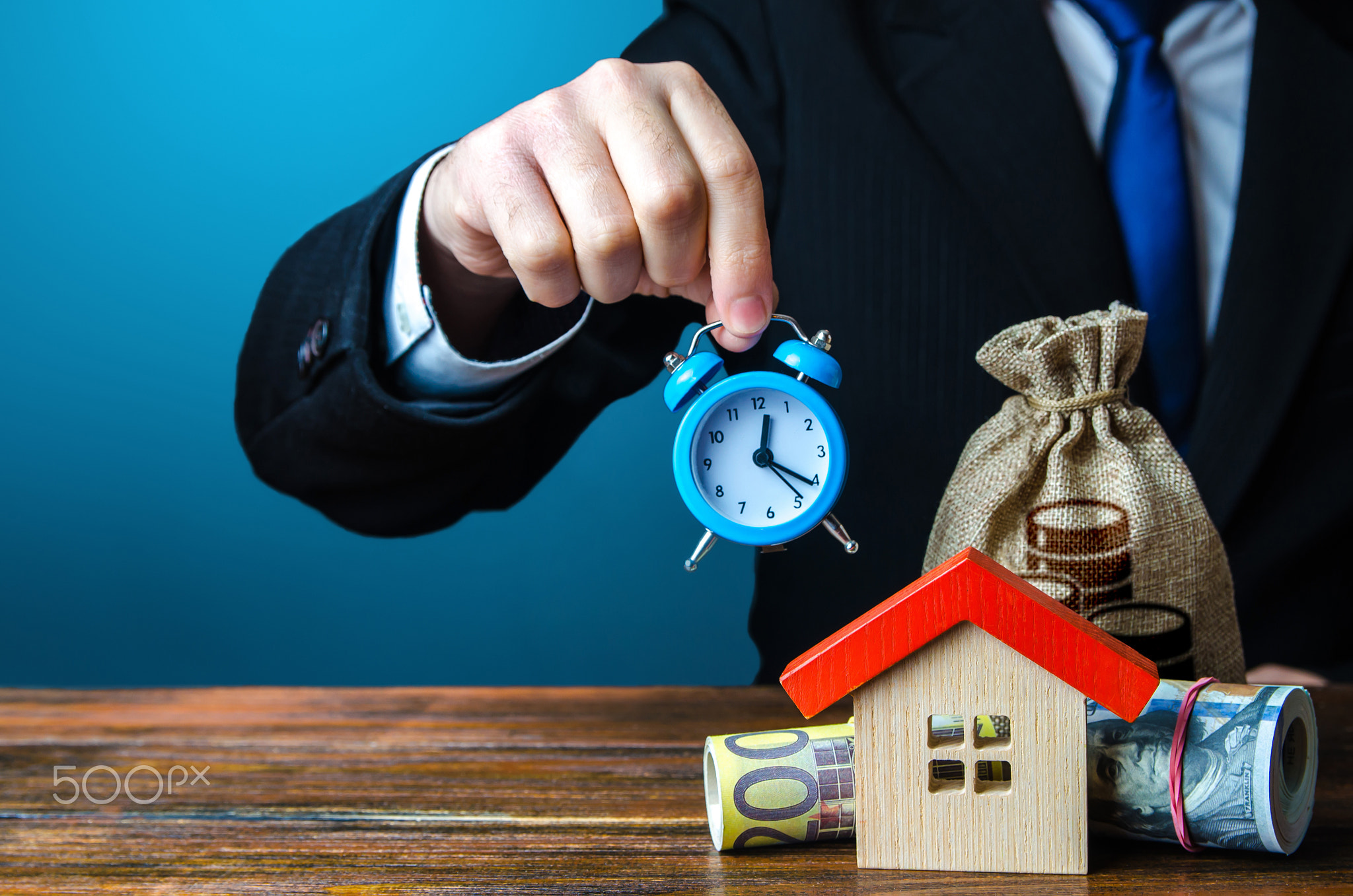 A man holds an alarm clock over the house and money.