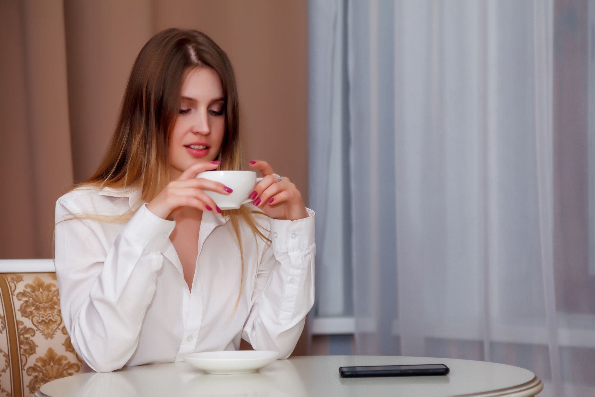 Girl of Slavic appearance with phone and white Cup is sitting at table