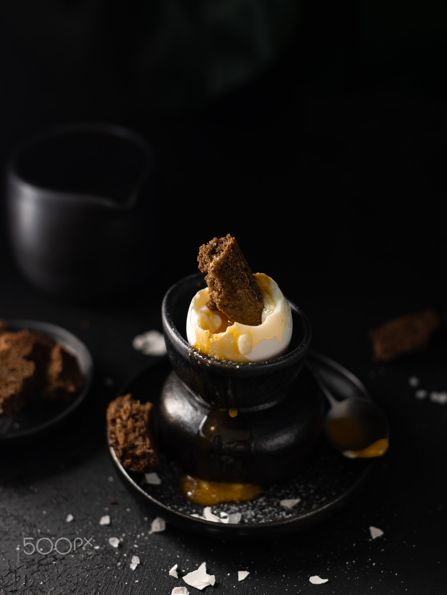 Boiled egg in a black ceramic stand with bread and salt on dark table