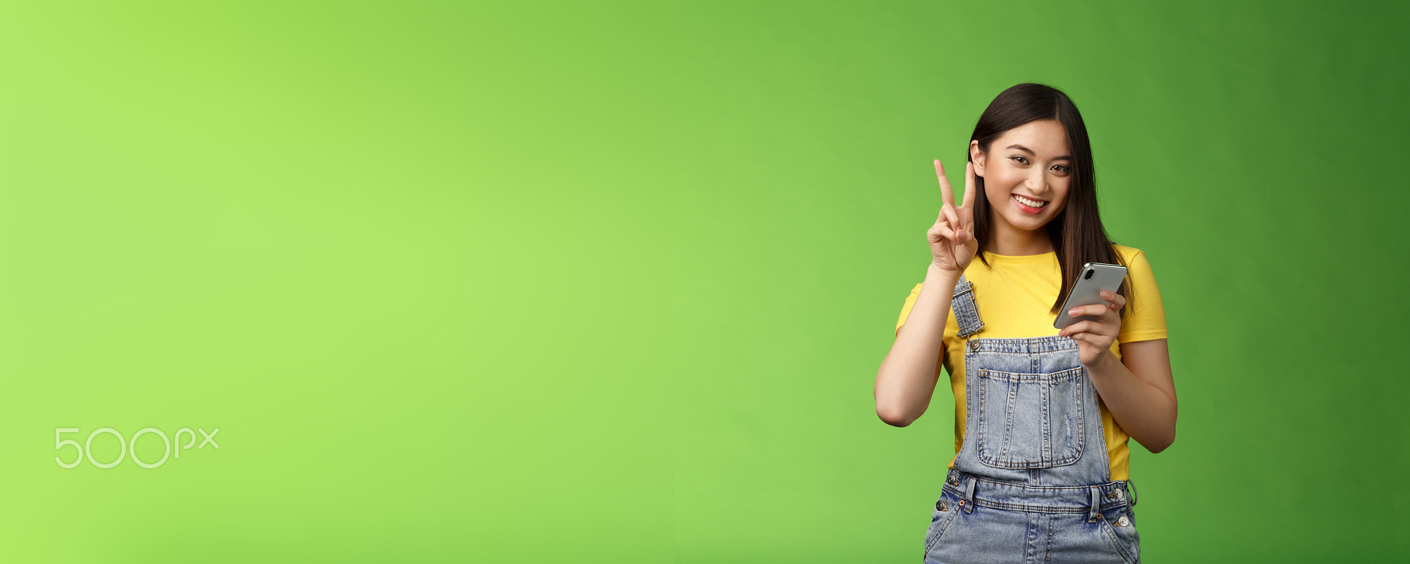 Cute tender asian woman hold smartphone, show victory peace sign, look