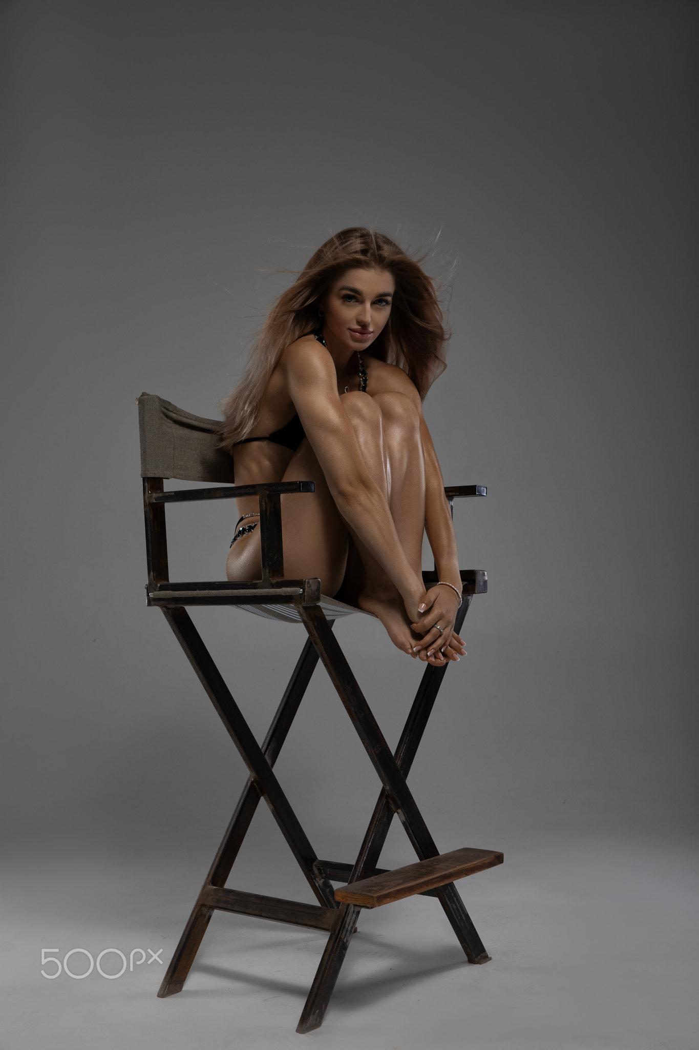 Girl in black swimsuit sits on a chair, raising up and huggin her legs