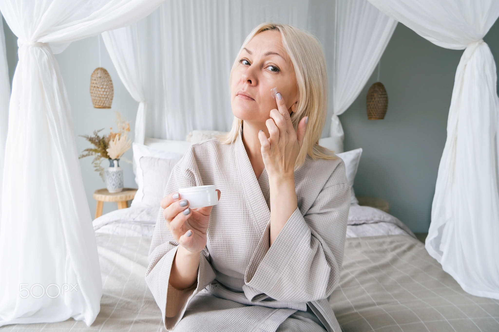 Middle aged blonde woman 50s takes care of skin. She is standing in