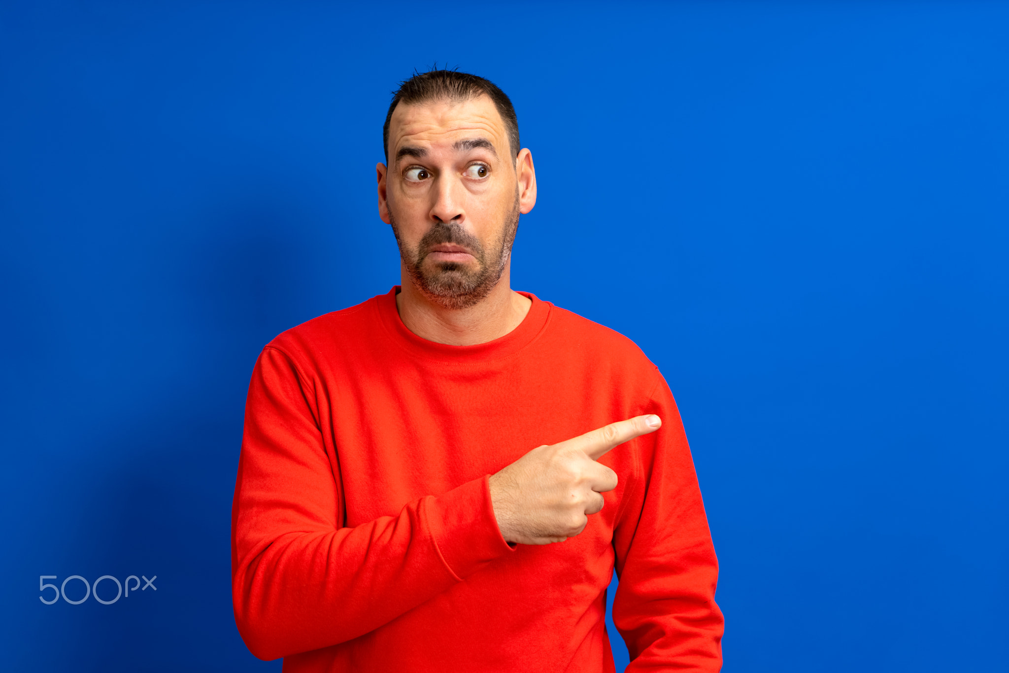 Bearded hispanic man dressed in red sweater pointing to the side with