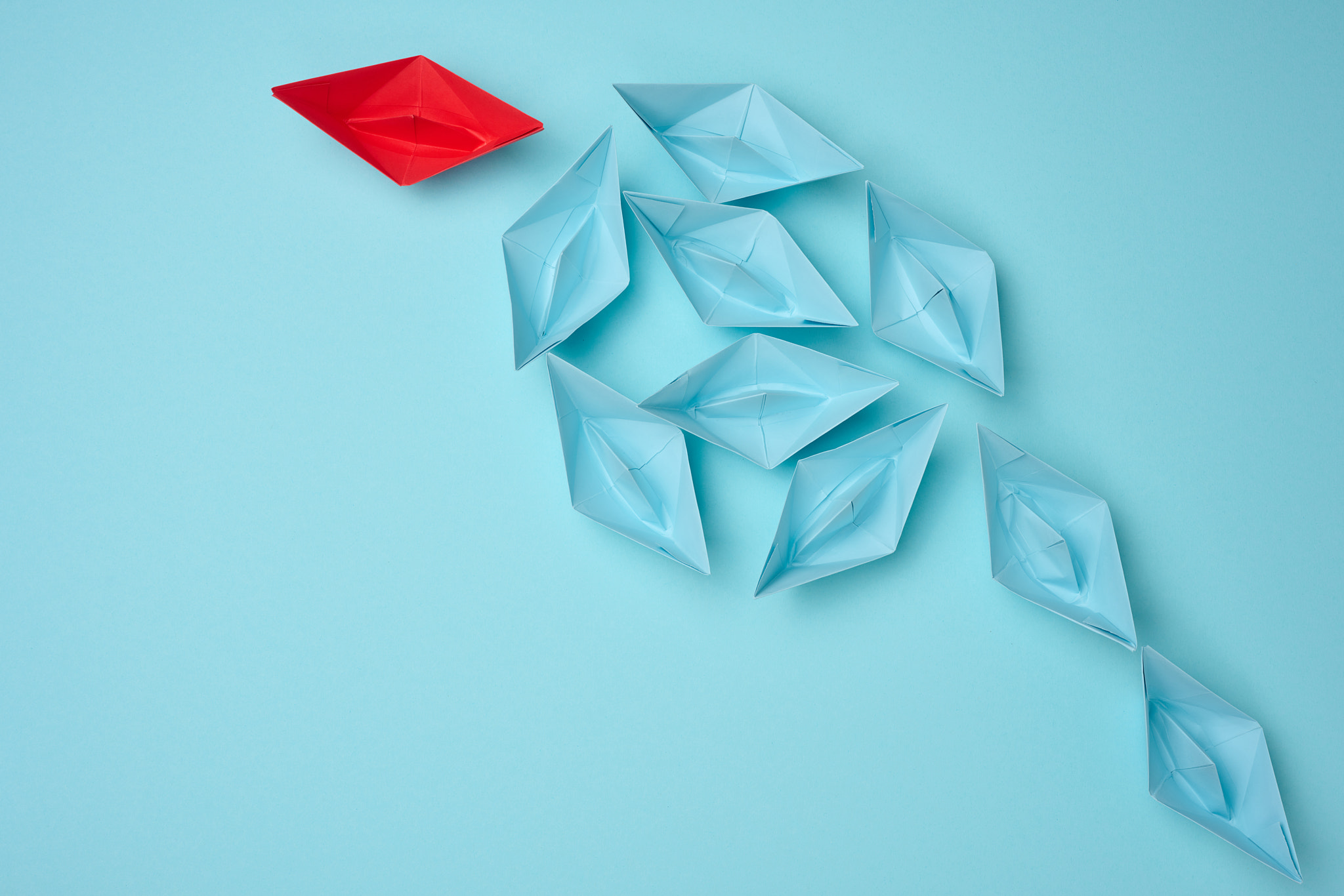 Group of blue paper boats heading in one direction and one red one heading 