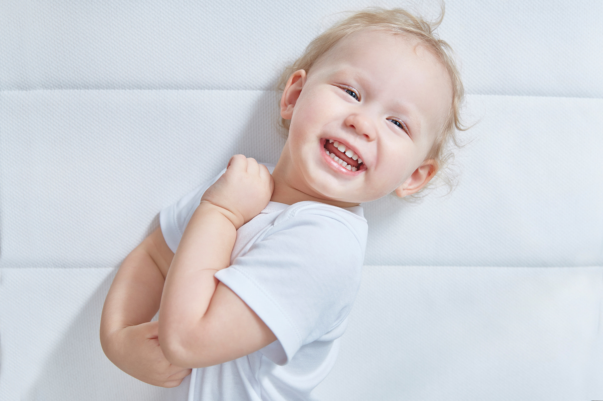 small child in white clothes is laughing happily lying on a white mattress in the bedroom