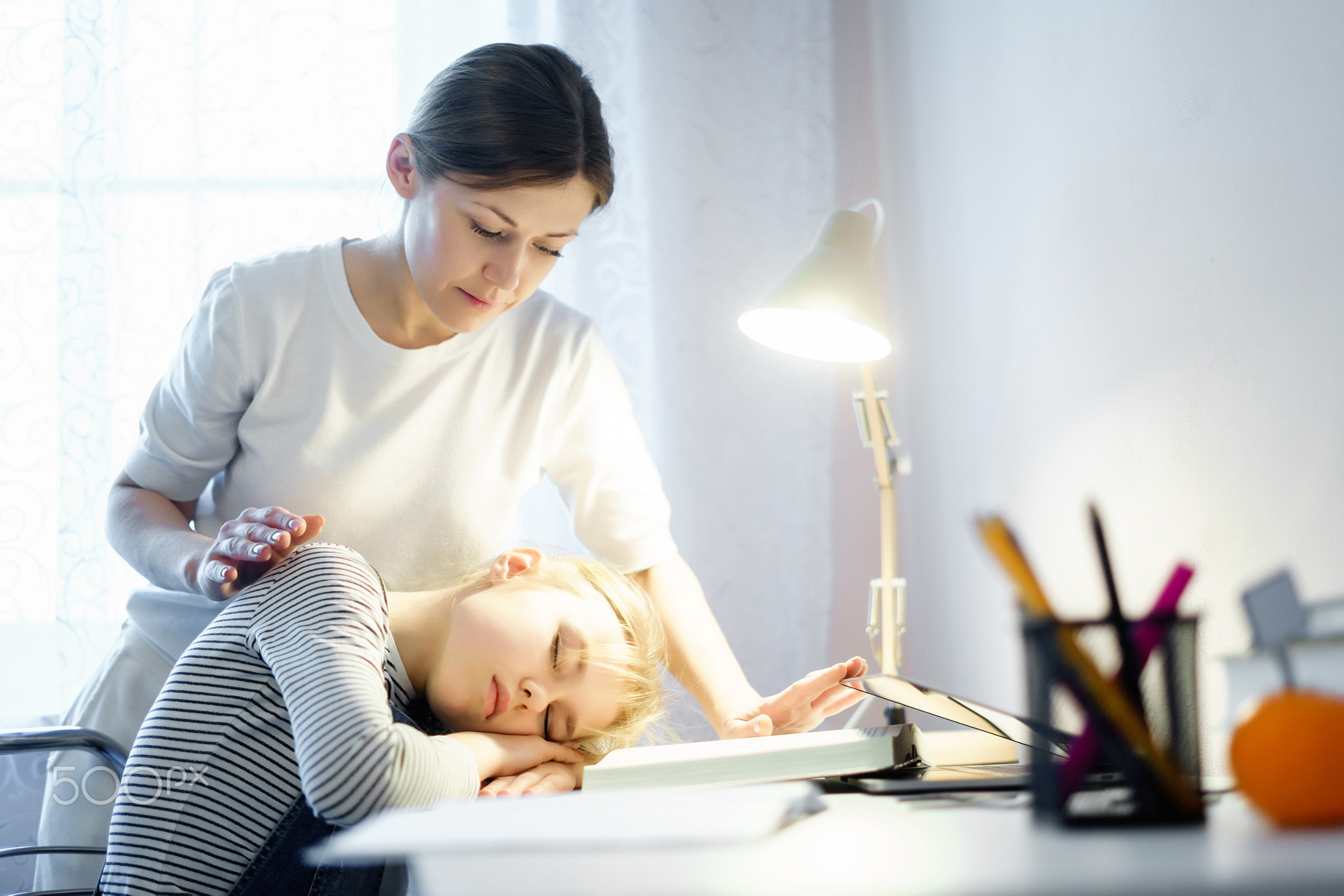 Mother standing near schoolgirl sleeping on books sitting at the table