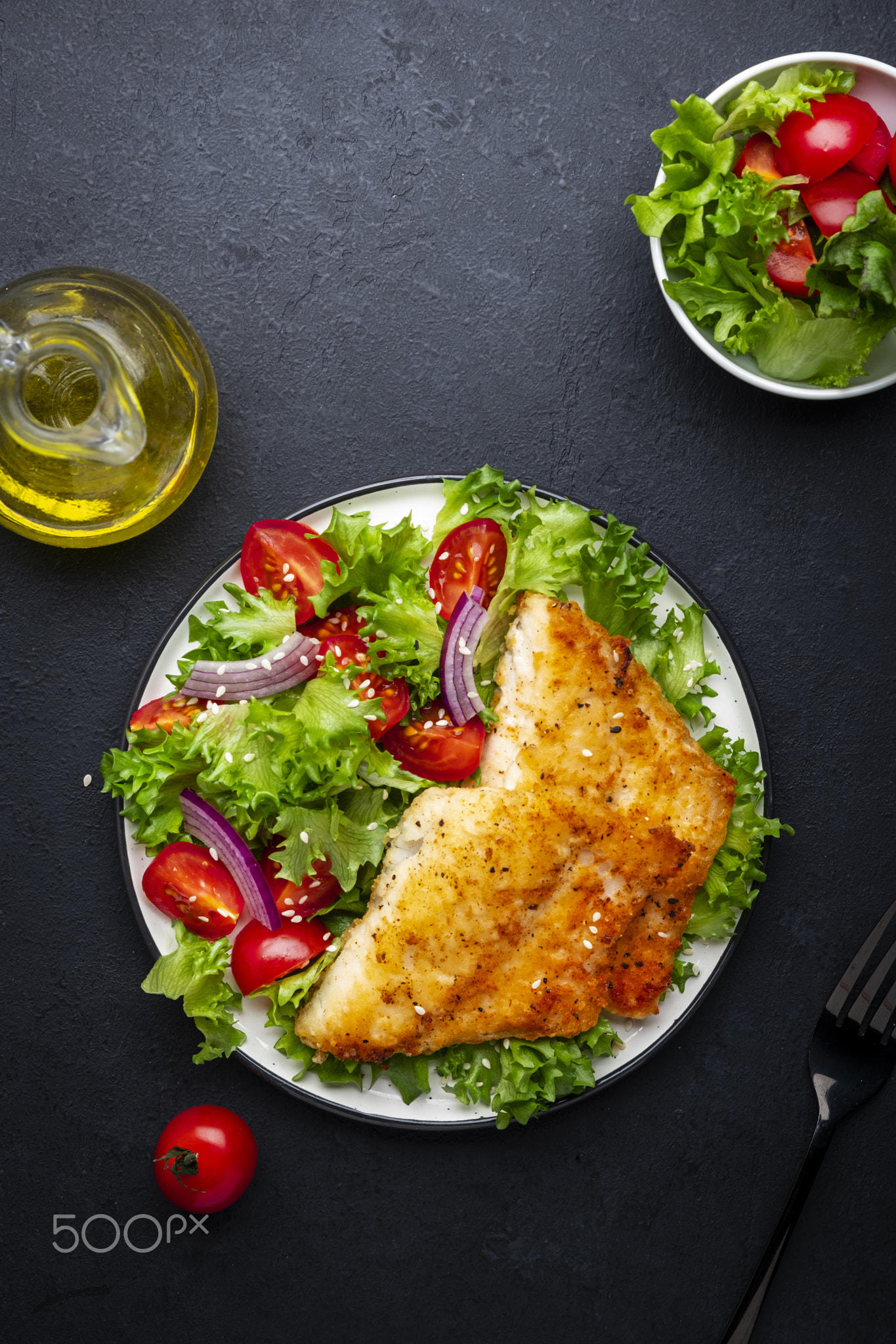 Fried cod fish with salad garnish from lettuce, cherry tomatoes and red onion with sesame seeds