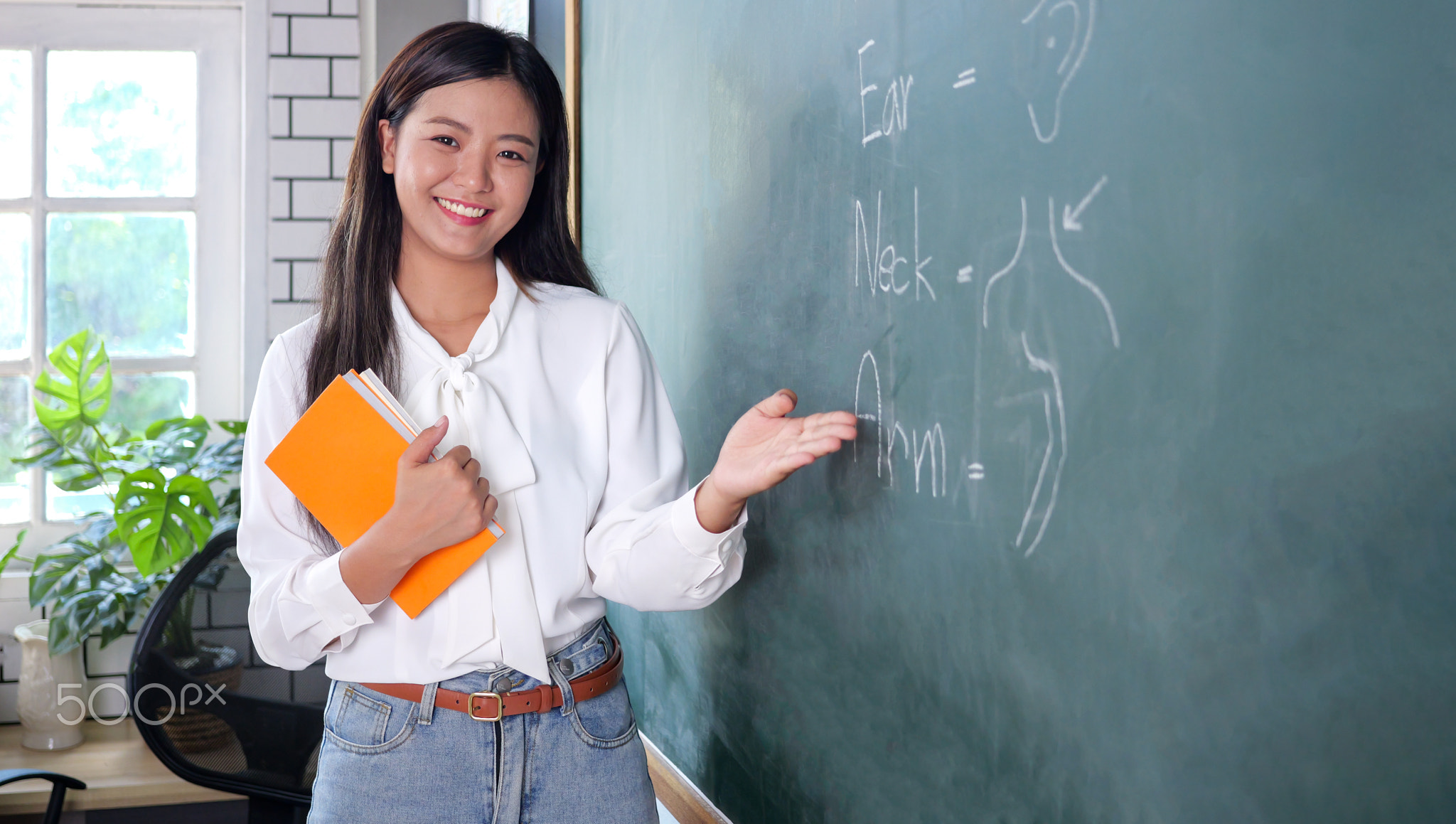 Teacher standing near backboard holding textbook posing to camera with