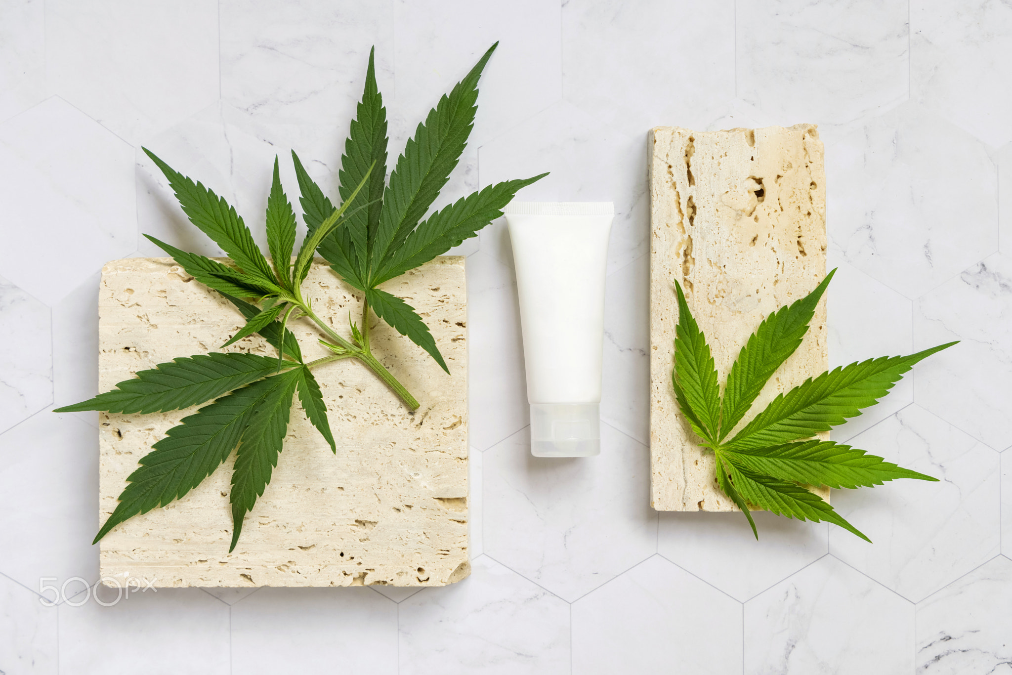 Cosmetic cream tube with blank label near green cannabis leaves and stones top view. Mockup