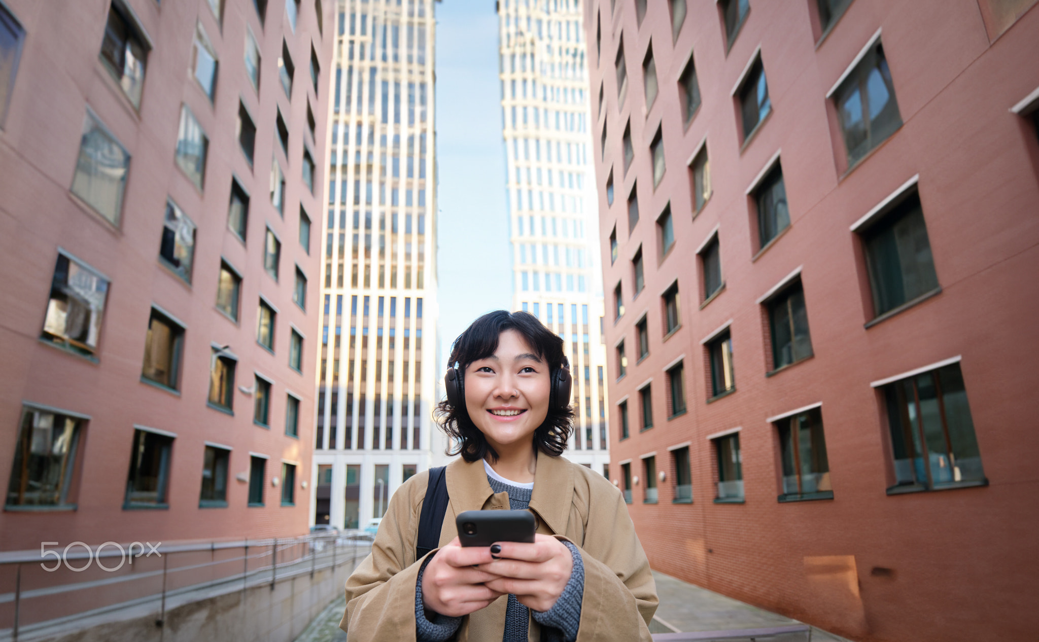 Portrait of cute girl tourist, korean woman in headphones, looks at mobile phone, uses smartphone