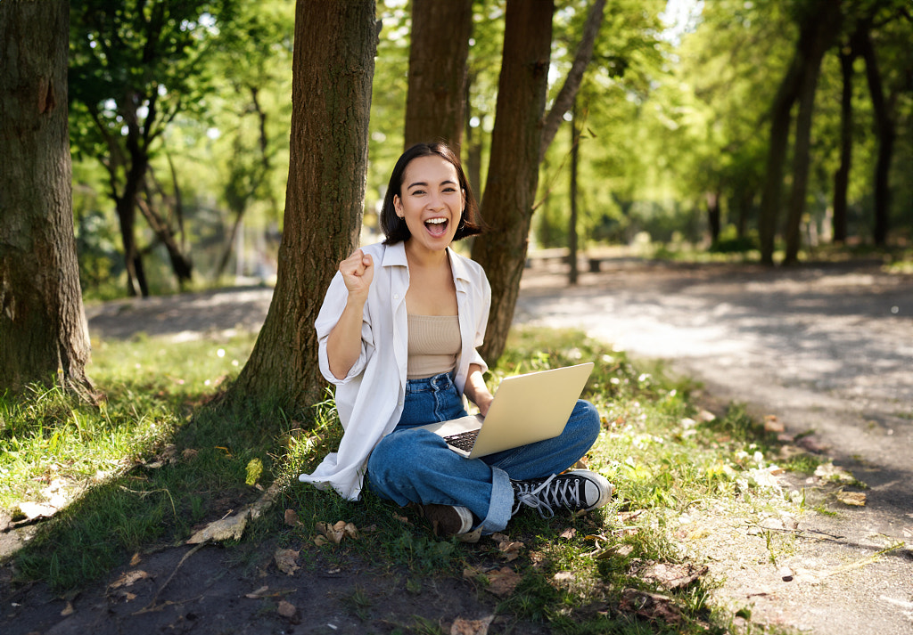 בחורה אסייתית צעירה ונלהבת, יושבת עם מחשב נייד ליד עץ בפארק שטוף שמש ירוק, חוגגת על ידי Mix and Match Studio ב-500px.com