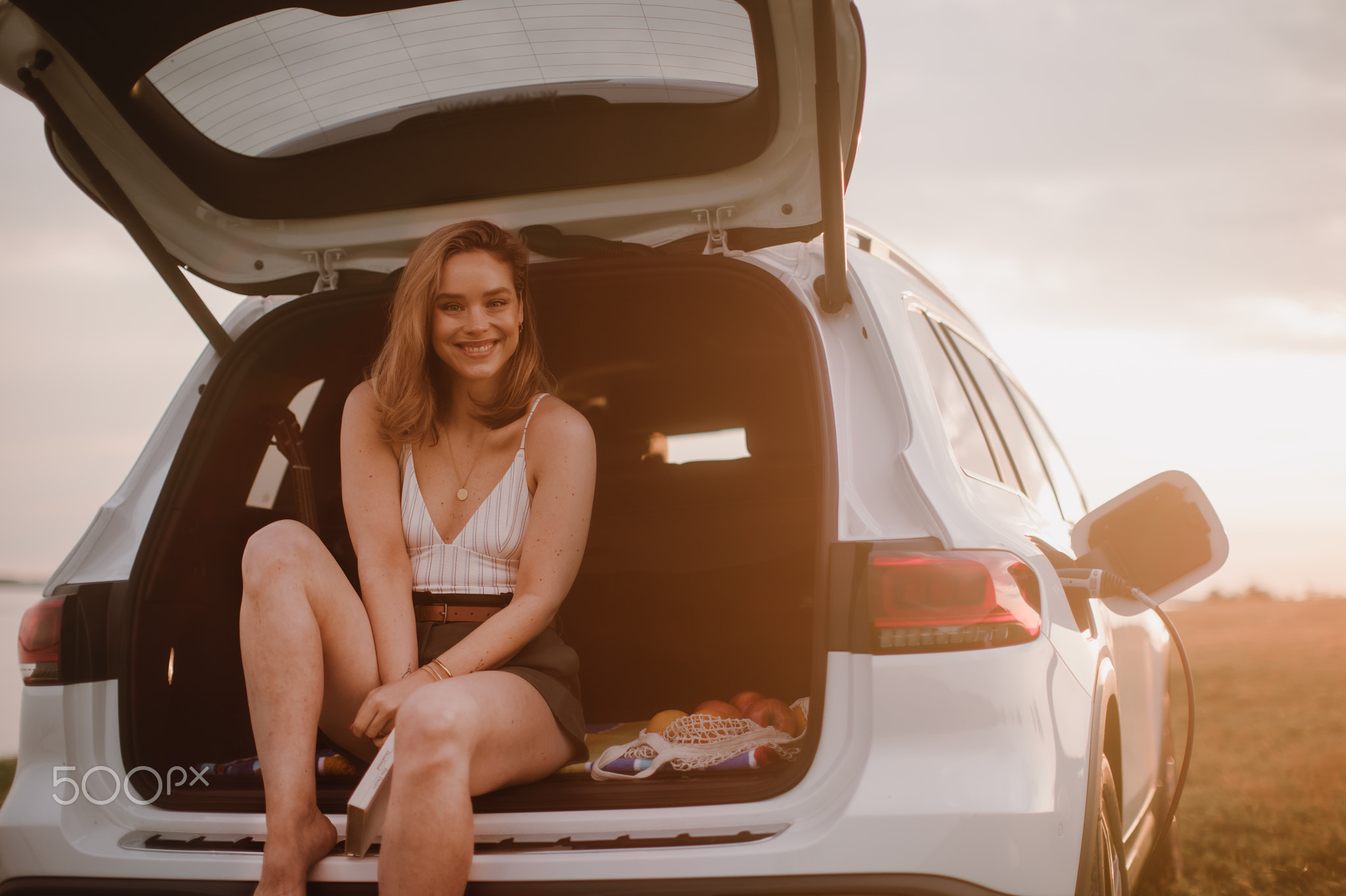 Young woman sitting in a car trunk, waiting for charging.