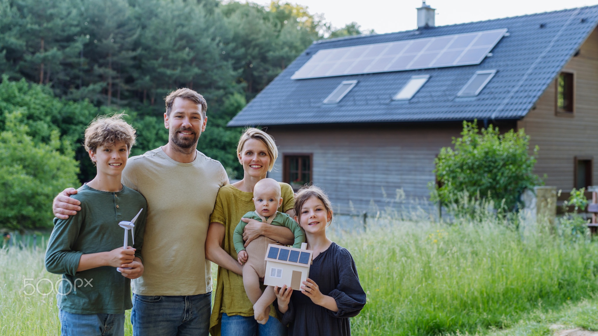 Happy family holding model of wind turbine and paper house with photovoltaics, standing near their