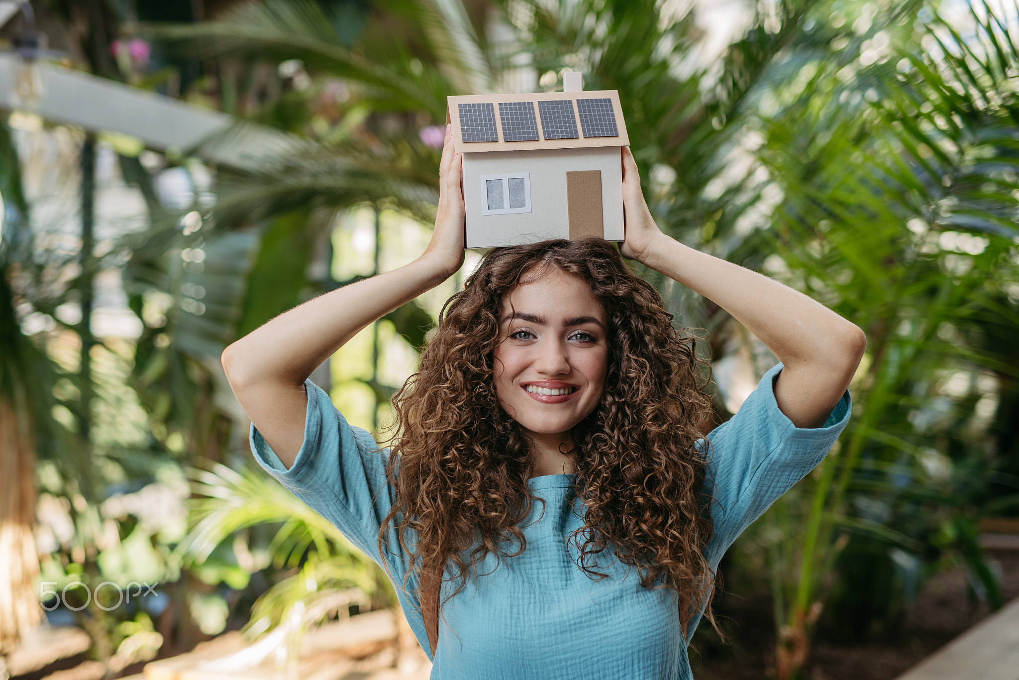 Young woman in botanical garden holding model of house with solar panels, concept of green energy.