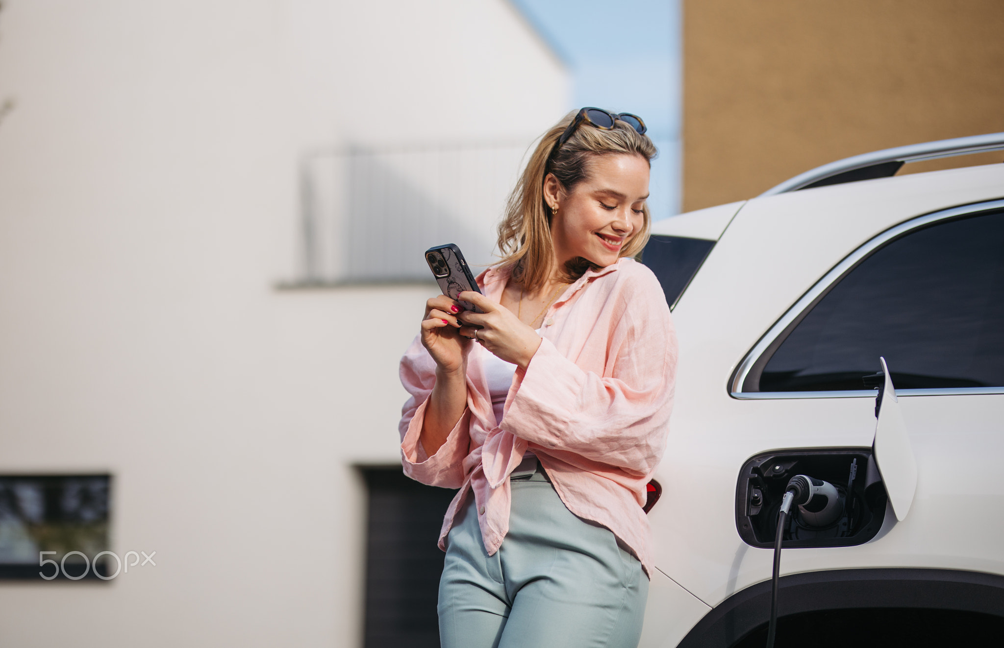 Young woman with smartphone charging her electric car in home, sustainable and economic