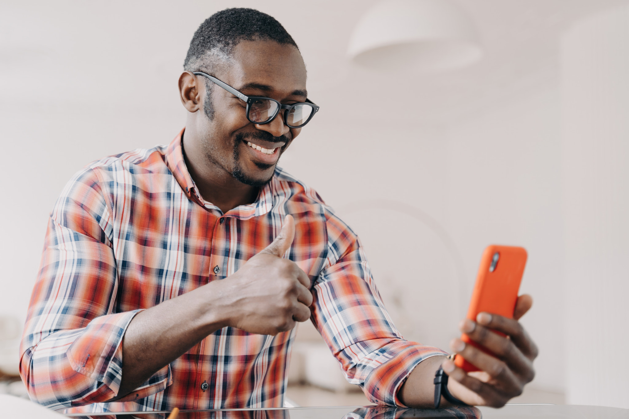 Smiling African-American man in glasses shows thumbs up, chats on video call, holds smartphone at de