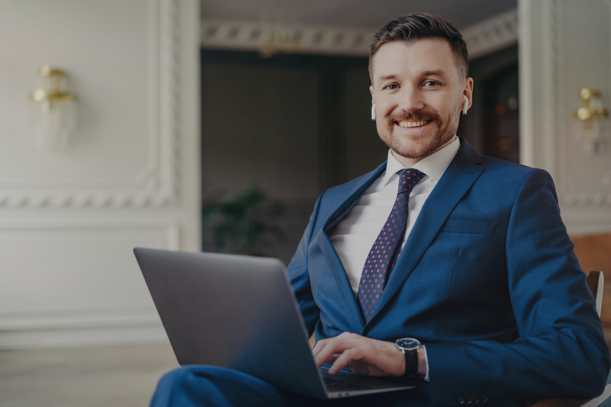 Confident young businessman in formal suit, working with laptop in office, smiling at camera, feelin